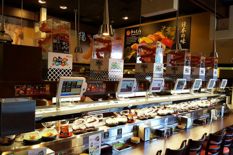 Bên trong một nhà hàng Kura Sushi, với băng chuyền sushi, bảng điện tử đặt món và băng tải chuyển thức ăn khác sushi ở trên.